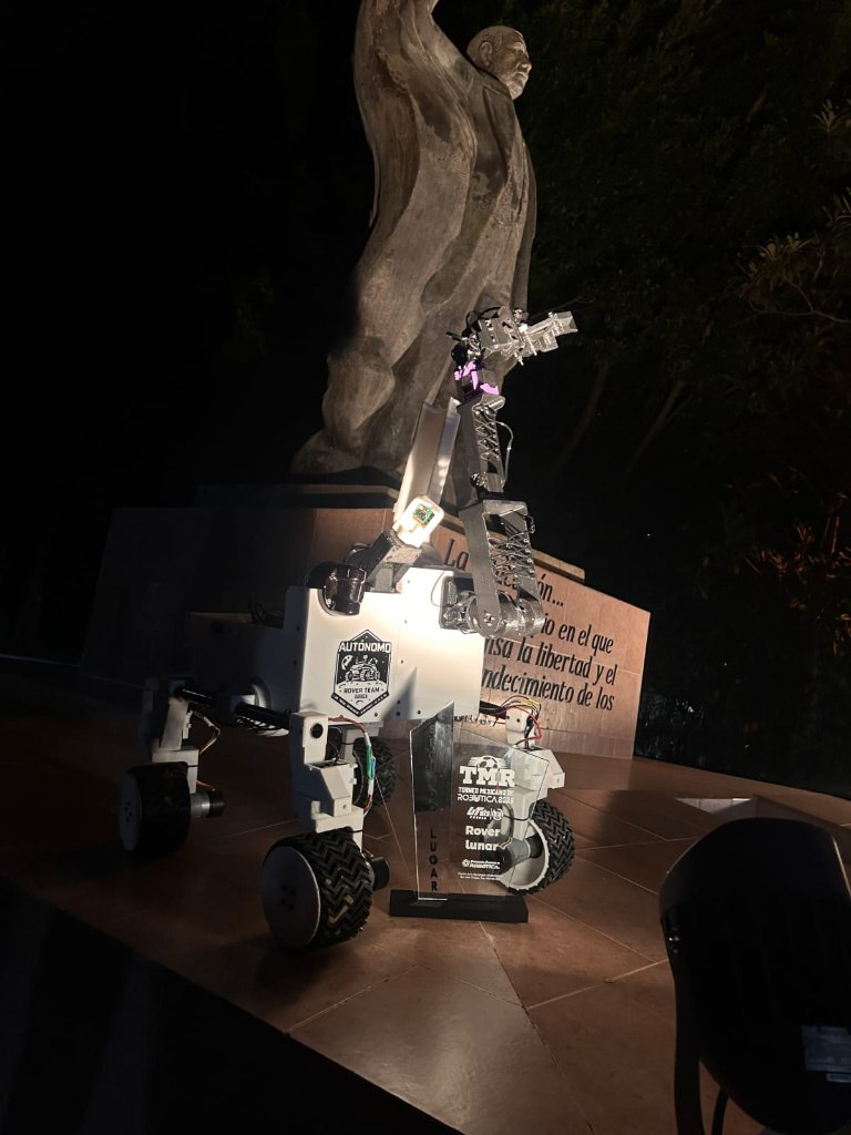 Rover AUTÓNOMO de noche frente al monumento de la UTM, con el trofeo del TMR 2026 al frente